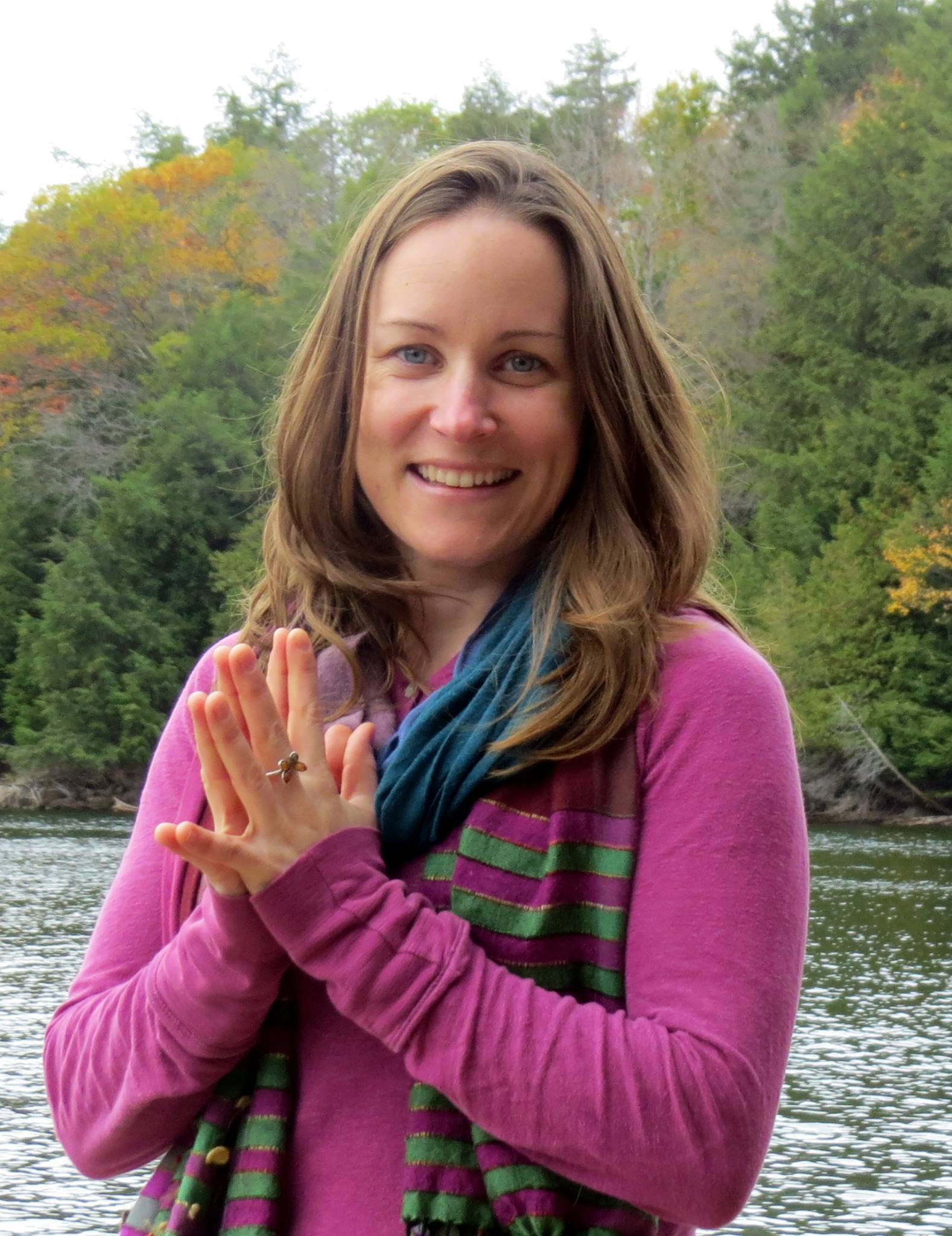 Shannon Crow, a white woman with brown hair, holds her hands in prayer position. She is wearing a purple blouse and a blue scarf, and is standing outdoors in front of a river and forest.