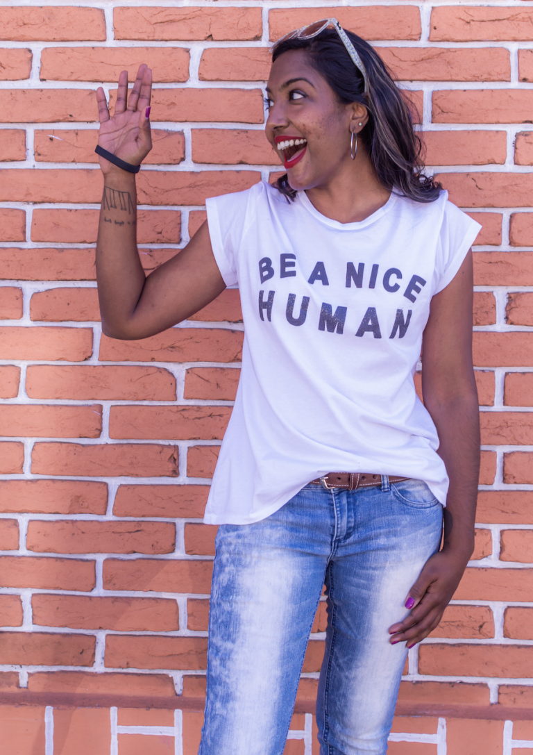 Crunch is standing in front of a red brick wall. She is looking to her right and is waving, She is wearing a white shirt that reads "Be a Nice Human" and blue jeans.